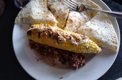 A plate with grilled corn on the cob, seasoned, and three quesadillas, with a side of beans.