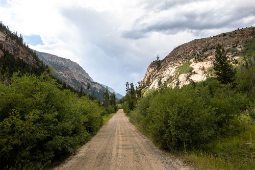 A dirt road stretches between lush greenery and rocky mountains under a cloudy sky.