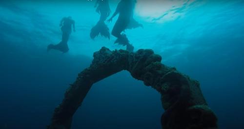Underwater scene featuring mermaids swimming near a large, ornate archway covered in marine life.