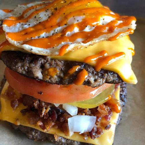 A stacked burger with two beef patties, cheese, bacon, tomato, pickles, onions, and a fried egg drizzled with sauce.