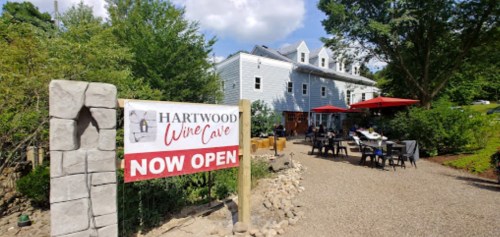 A sign reading "Hartwood Wine Cave Now Open" in front of a charming building with outdoor seating and greenery.