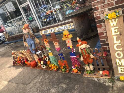 Colorful scarecrows and autumn decorations line a sidewalk outside a flower shop, welcoming the fall season.
