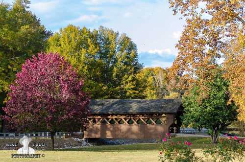 A scenic view of a covered bridge surrounded by colorful autumn trees and a grassy area.