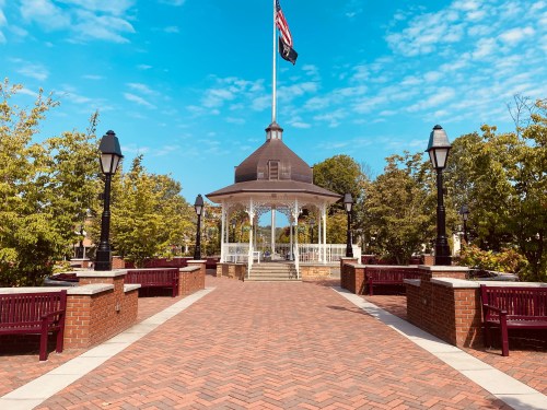 A brick pathway leads to a gazebo with an American flag, surrounded by trees and benches under a blue sky.