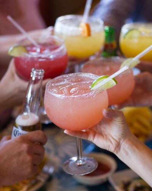 A group of hands holding colorful cocktails and drinks with garnishes, celebrating together at a table.