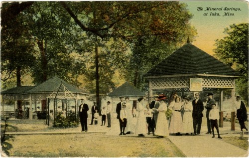 A vintage postcard depicting a group of people in white attire at the Mineral Springs in Iuka, Mississippi.