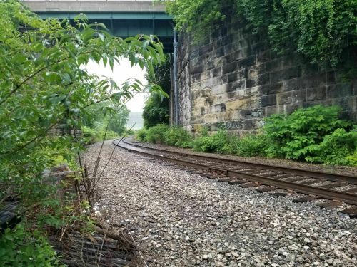 A view of a railway track curving under a bridge, surrounded by greenery and stone walls.