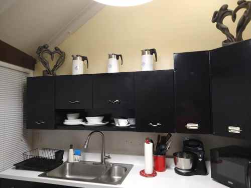 A kitchen with black cabinets, white dishes, and several coffee pots on top. A sink and countertop are visible.