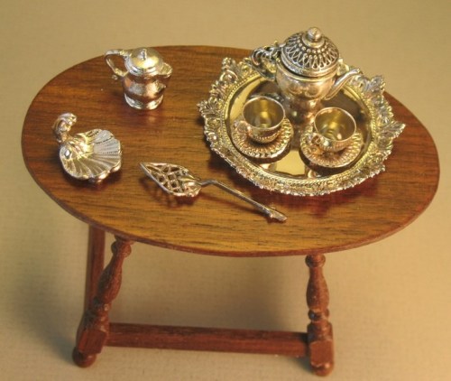 A miniature wooden table displaying a silver tea set, including a teapot, cups, and a serving tray.