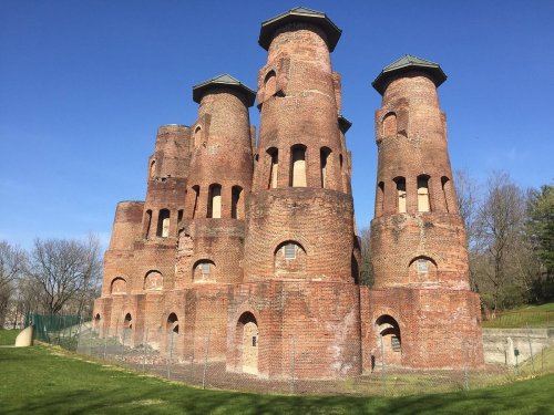 Historic brick structure with multiple towers against a clear blue sky, surrounded by greenery.
