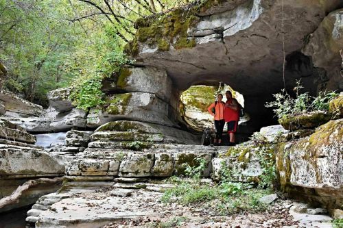Two people and a dog stand under a large rock formation in a lush, green forested area.