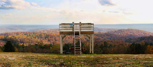 A wooden observation deck overlooks a colorful autumn landscape with rolling hills and a cloudy sky.