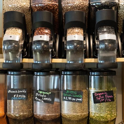 Dispensers of various lentils and split peas, labeled with prices and organic tags, arranged on a wooden shelf.
