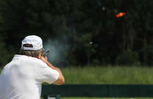 A person in a white cap aims a shotgun at a flying target in a green outdoor setting.