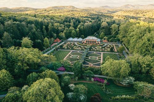 Aerial view of a lush garden with geometric flower beds, surrounded by trees and rolling hills in the background.