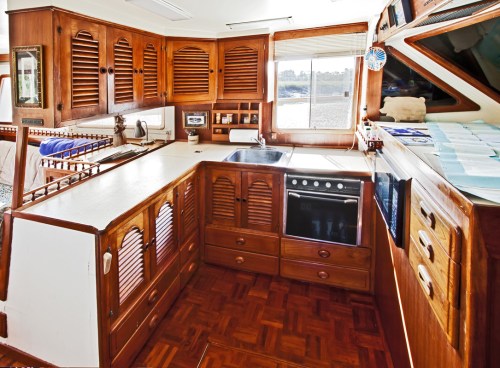 A wooden kitchen with louvered cabinets, a sink, oven, and a window showing a view outside. Bright and spacious design.