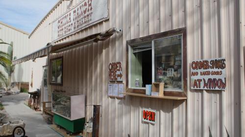 A rustic bakery with a window labeled "Open" and signs for ordering baked goods. Palm trees in the background.