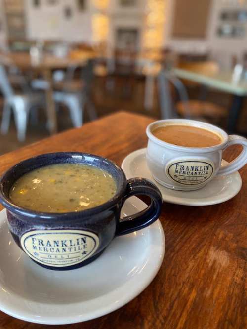 Two bowls of soup on white plates, one dark and one light, on a wooden table in a cozy café setting.