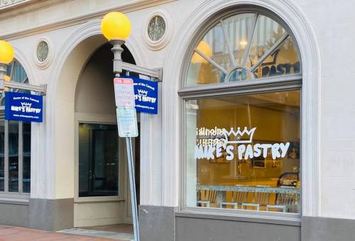 Exterior of Mike's Pastry shop, featuring large windows and a sign above the entrance. Street signs visible nearby.