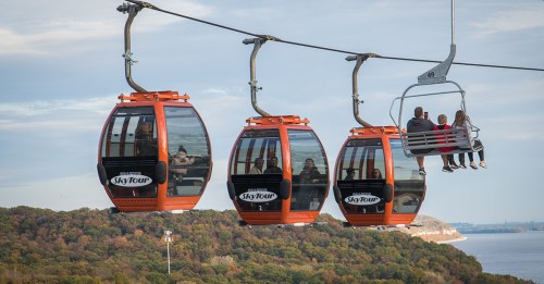 Three orange gondolas on a cable line, with a chairlift carrying passengers in the background against a scenic landscape.