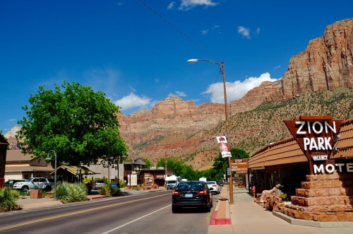 A scenic view of a street in Zion National Park, featuring mountains, trees, and a motel sign under a blue sky.