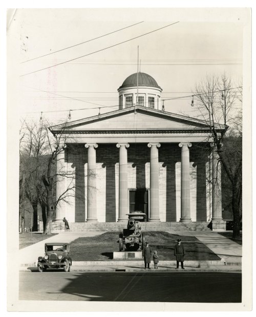 Historic building with a dome and columns, featuring a vintage car and two people in front, surrounded by trees.