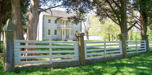 A historic two-story house behind a white wooden fence, surrounded by trees and a grassy area.