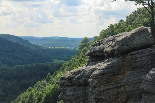 A scenic view of rolling hills and valleys, with a rocky outcrop in the foreground and a cloudy sky above.
