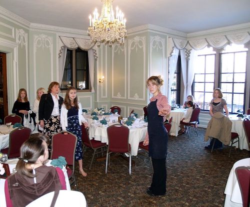 A group of women and girls in a decorated dining room with tables set for an event, featuring a chandelier above.