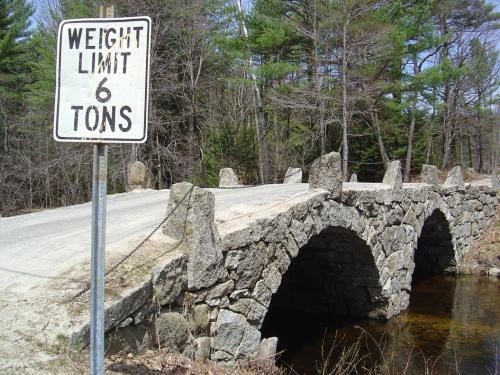 A stone arch bridge with a sign indicating a weight limit of 6 tons, surrounded by trees and a small stream.