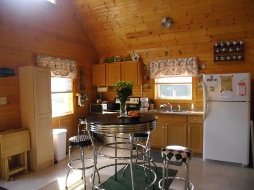 Cozy kitchen with wooden walls, a round table, bar stools, and appliances, featuring a sunny window and floral decor.
