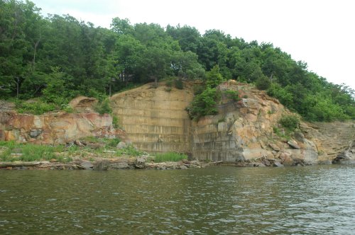 A rocky cliff with greenery on top, rising above a calm body of water under a cloudy sky.