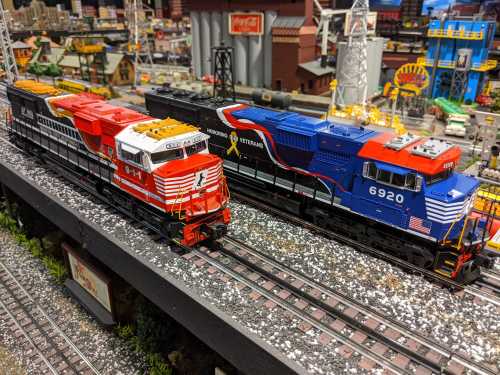 Two model trains on a track, one red and white, the other blue and red, set in a detailed miniature landscape.