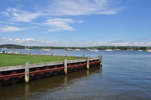 A calm waterfront scene with sailboats anchored in the distance under a blue sky with scattered clouds.