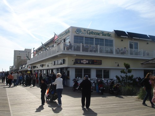A busy boardwalk scene featuring the Greene Turtle restaurant, with people walking and enjoying the sunny day.