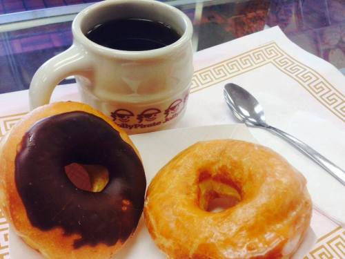 A cup of black coffee beside two donuts: one chocolate-glazed and one glazed, on a white napkin with a spoon.
