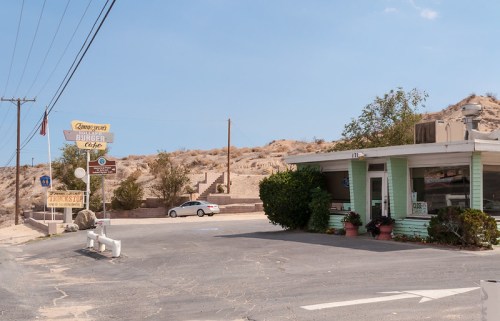 A small, retro diner with green trim, surrounded by desert landscape and a clear blue sky.