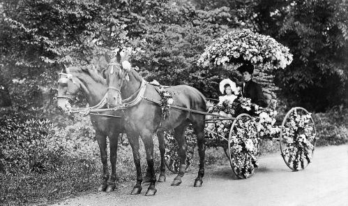 A vintage horse-drawn carriage adorned with flowers, carrying a woman and a child through a lush, green landscape.