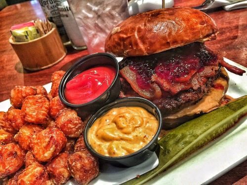 A juicy burger with bacon and sauce, served with tater tots, ketchup, and mustard on a plate with a pickle.