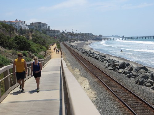Two people walk along a coastal path beside a railway track, with waves crashing on the beach and palm trees in the background.