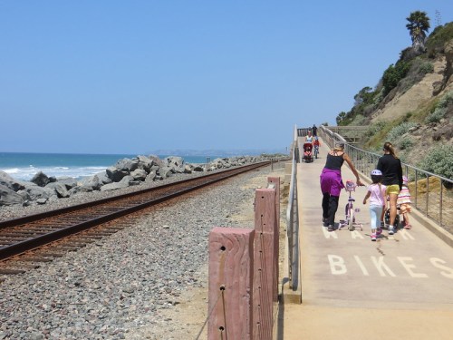 A scenic coastal path with people walking and biking alongside the ocean and railway tracks.
