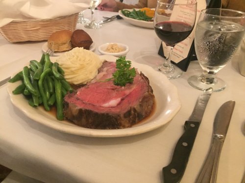 A plate of prime rib with green beans, mashed potatoes, and rolls, accompanied by red wine and water on a dining table.