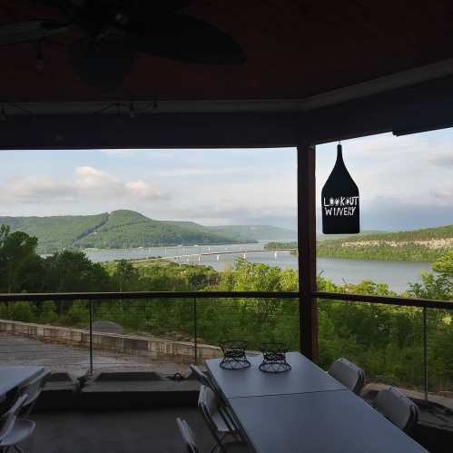 Scenic view from a winery deck overlooking a river and lush green hills under a cloudy sky.