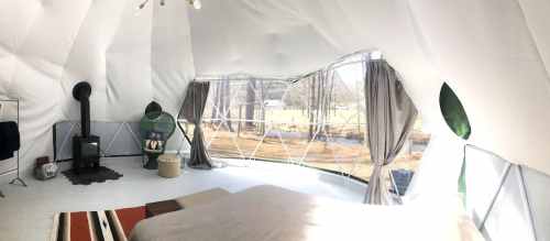 Interior of a geodesic dome tent featuring a bed, wood stove, and large windows with a view of nature outside.