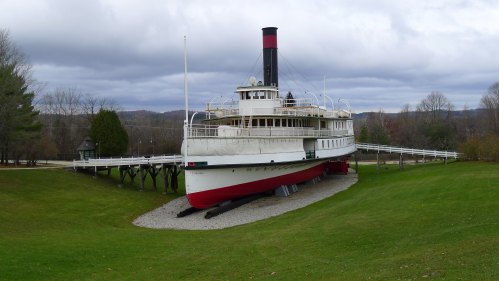 A historic steamboat on land, surrounded by grass and trees, under a cloudy sky.