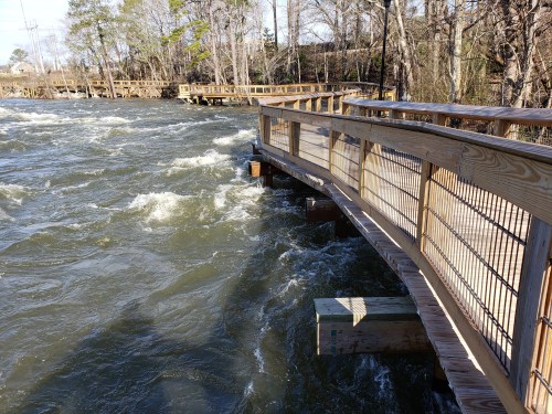 A wooden walkway curves over turbulent water, surrounded by trees and a natural landscape.