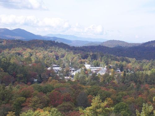 A scenic view of a small town surrounded by vibrant autumn foliage and distant mountains under a partly cloudy sky.