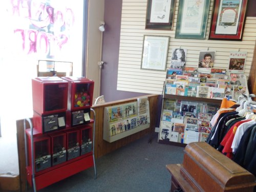 Interior of a vintage store featuring vending machines, magazines, and clothing racks against a wall with framed art.