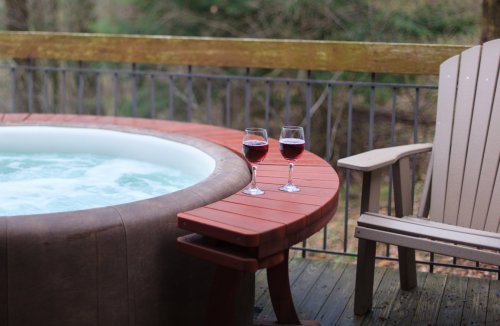 Two glasses of red wine sit on a wooden table next to a hot tub on a deck surrounded by trees.