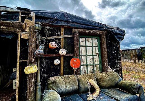 An abandoned, weathered cabin with a dark tarp, a worn couch, and Halloween decorations under a cloudy sky.